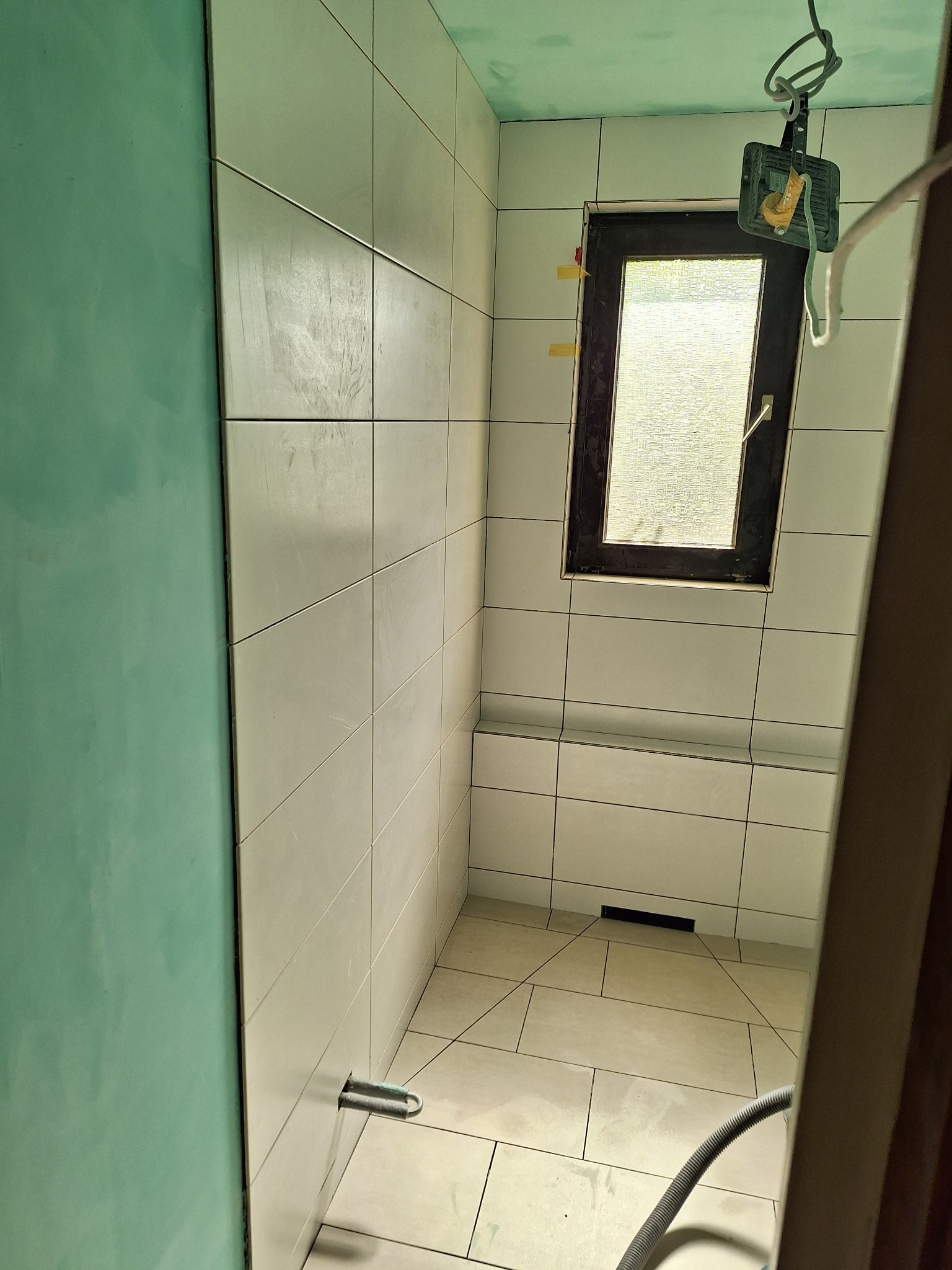 Wnętrze łazienki w trakcie prac wykończeniowych: białe płytki na ścianach, okno z ciemną ramą, fragment zielonej ściany i sufitu, widoczne narzędzia i oświetlenie budowlane.