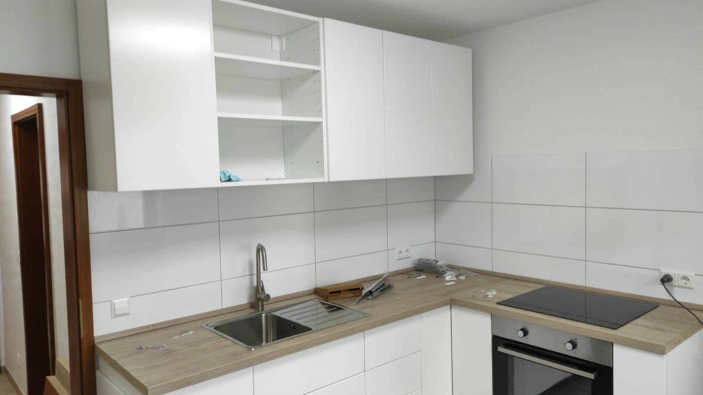 Montaż białych szafek kuchennych z drewnianym blatem i płytkami na ścianie, widoczny zlew z kranem oraz płyta indukcyjna nad piekarnikiem, narzędzia na blacie, wnętrze w trakcie prac.