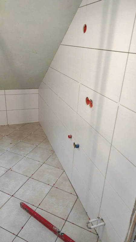 Ściana w skosie wyłożona białymi płytkami ceramicznymi z otworami na instalacje, podłoga w jasnych płytkach, czerwona poziomica leżąca przy ścianie, widoczne krzyżyki dystansowe.