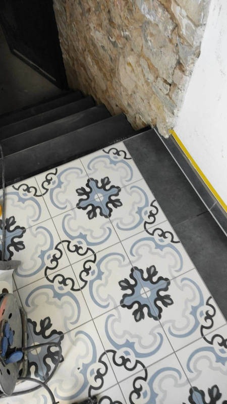 Wzorzyste płytki podłogowe w odcieniach błękitu i szarości, ułożone przy schodach z ciemnymi stopnicami i kamienną ścianą, wykończone listwą przypodłogową w kolorze żółtym.