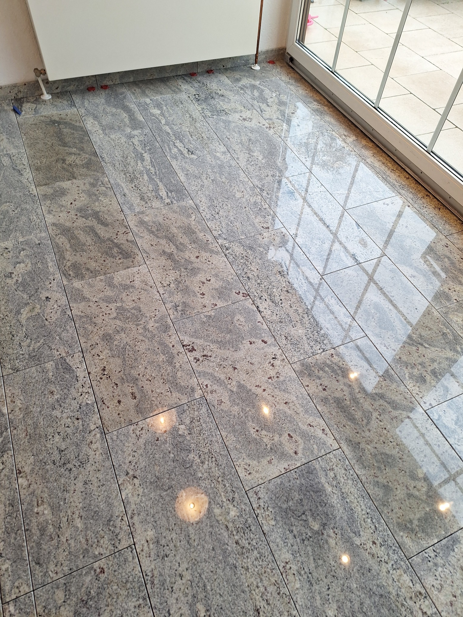 Ułożona podłoga z płytek imitujących kamień, widoczne krzyżyki dystansowe przy ścianie i odbicie okna w połyskującej powierzchni.