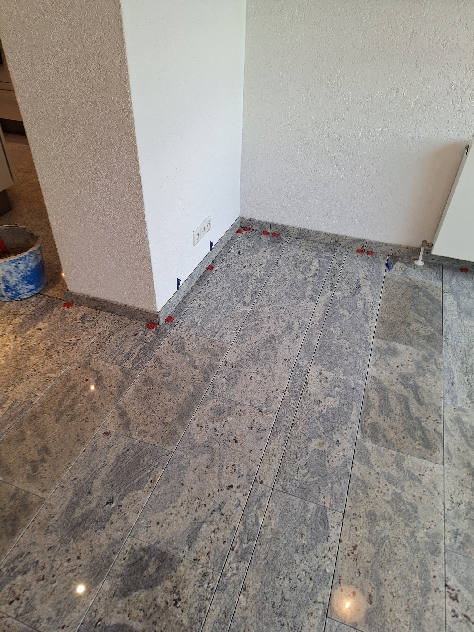 Układanie szarych płytek podłogowych z użyciem poziomujących klipsów i klinów w narożniku pomieszczenia w Głogowie. Widoczny fragment ściany z gniazdkami elektrycznymi.