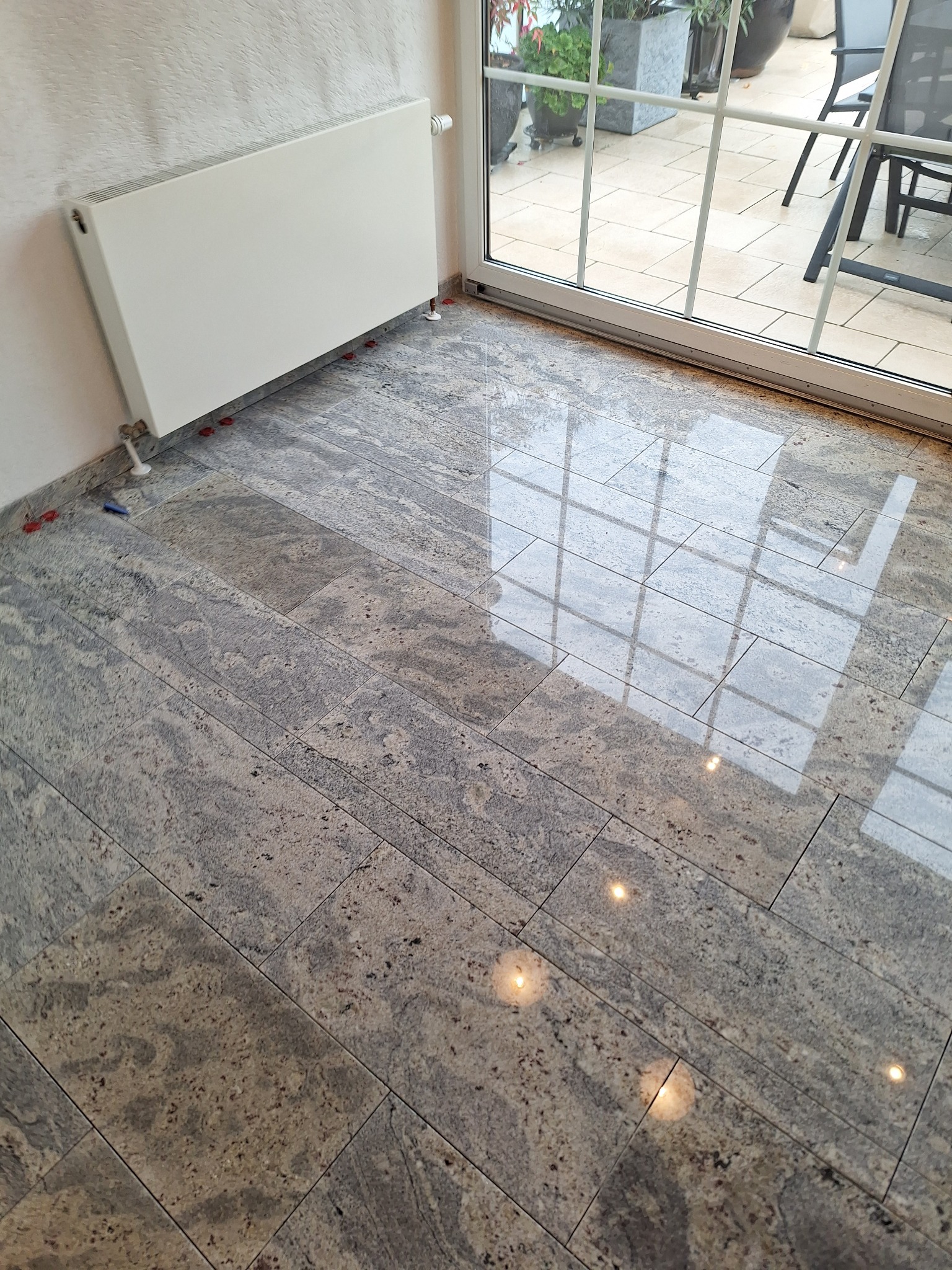 Ułożona, polerowana podłoga z płytek imitujących kamień, z widocznymi plastikowymi krzyżykami dystansowymi, grzejnik ścienny i odbicie światła w oknie.