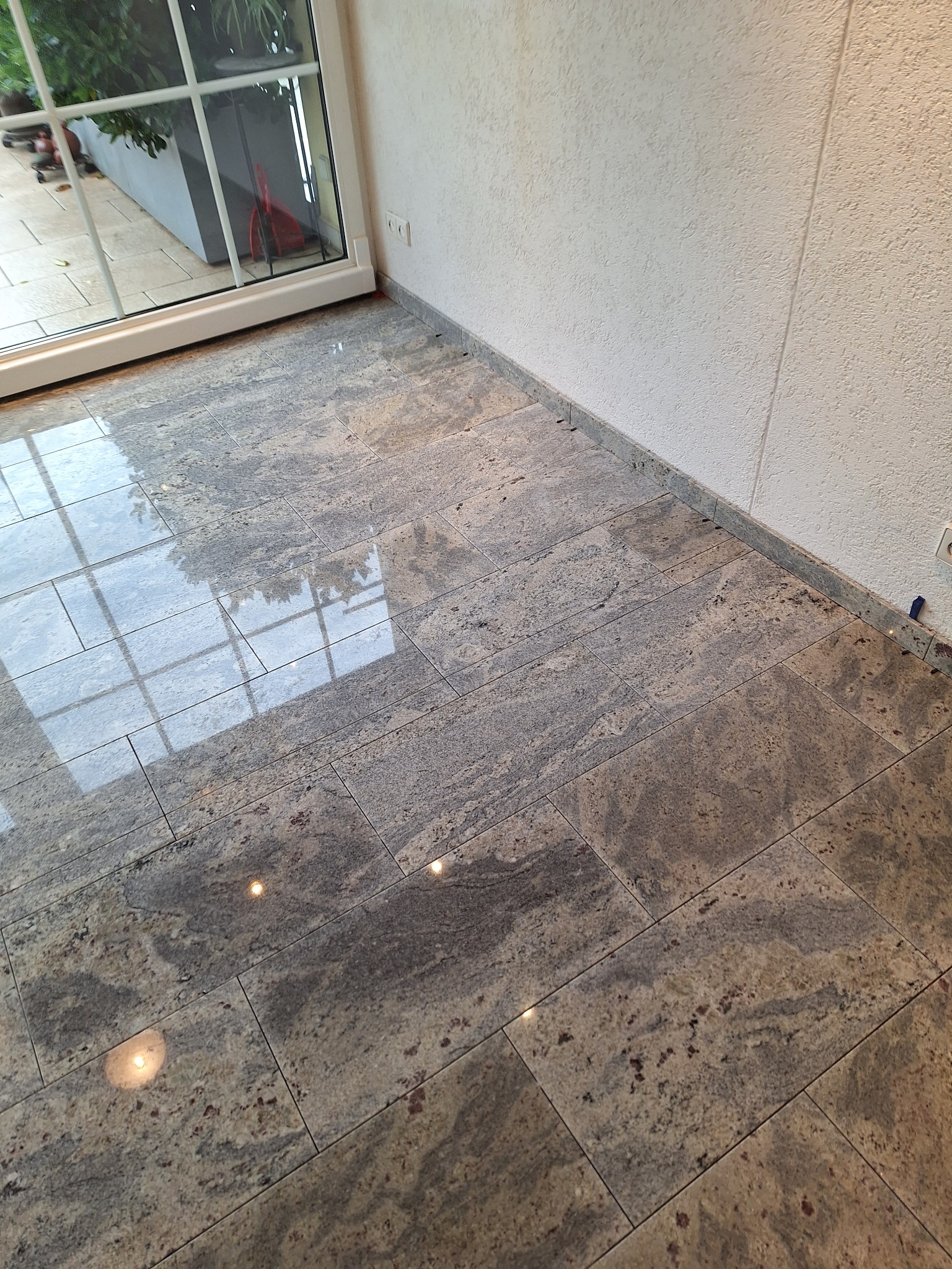 Ułożona podłoga z płytek imitujących kamień, z widocznym odbiciem okna i roślinności, oraz fragmentem ściany z fakturą baranka.