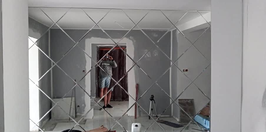Ściana wyłożona dekoracyjnymi lustrami w kształcie rombów, odbijającymi postać fotografa w pomieszczeniu w trakcie wykończenia, widoczne ślady szpachlowania na ścianach.