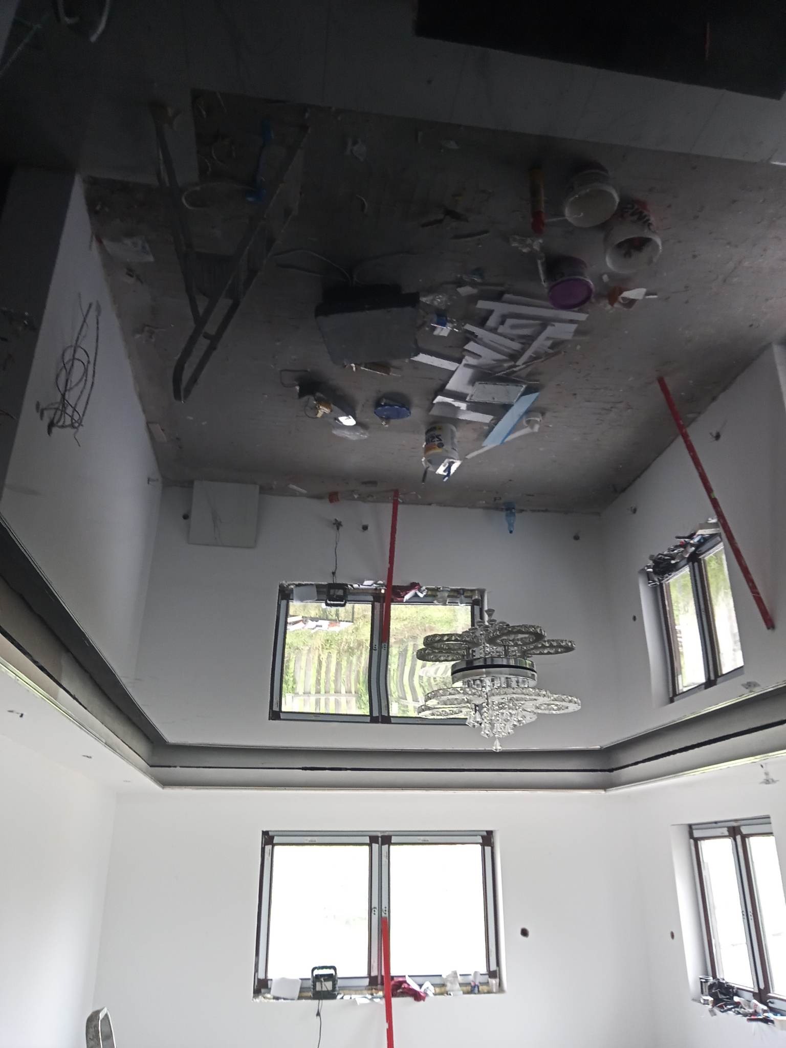 Pokój w trakcie remontu, widoczny surowy sufit z elementami instalacji, ściany po szpachlowaniu, zamontowane okna, wiszący kryształowy żyrandol. Na suficie widoczne puszki z farbą i narzędzia.