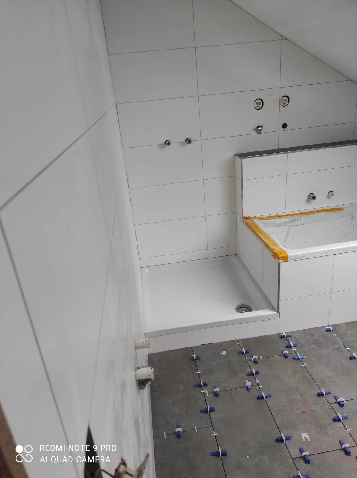 Biała łazienka w trakcie remontu z widocznym brodzikiem, wanną zabezpieczoną folią i nieukończoną podłogą z niebieskimi krzyżykami dystansowymi do płytek.