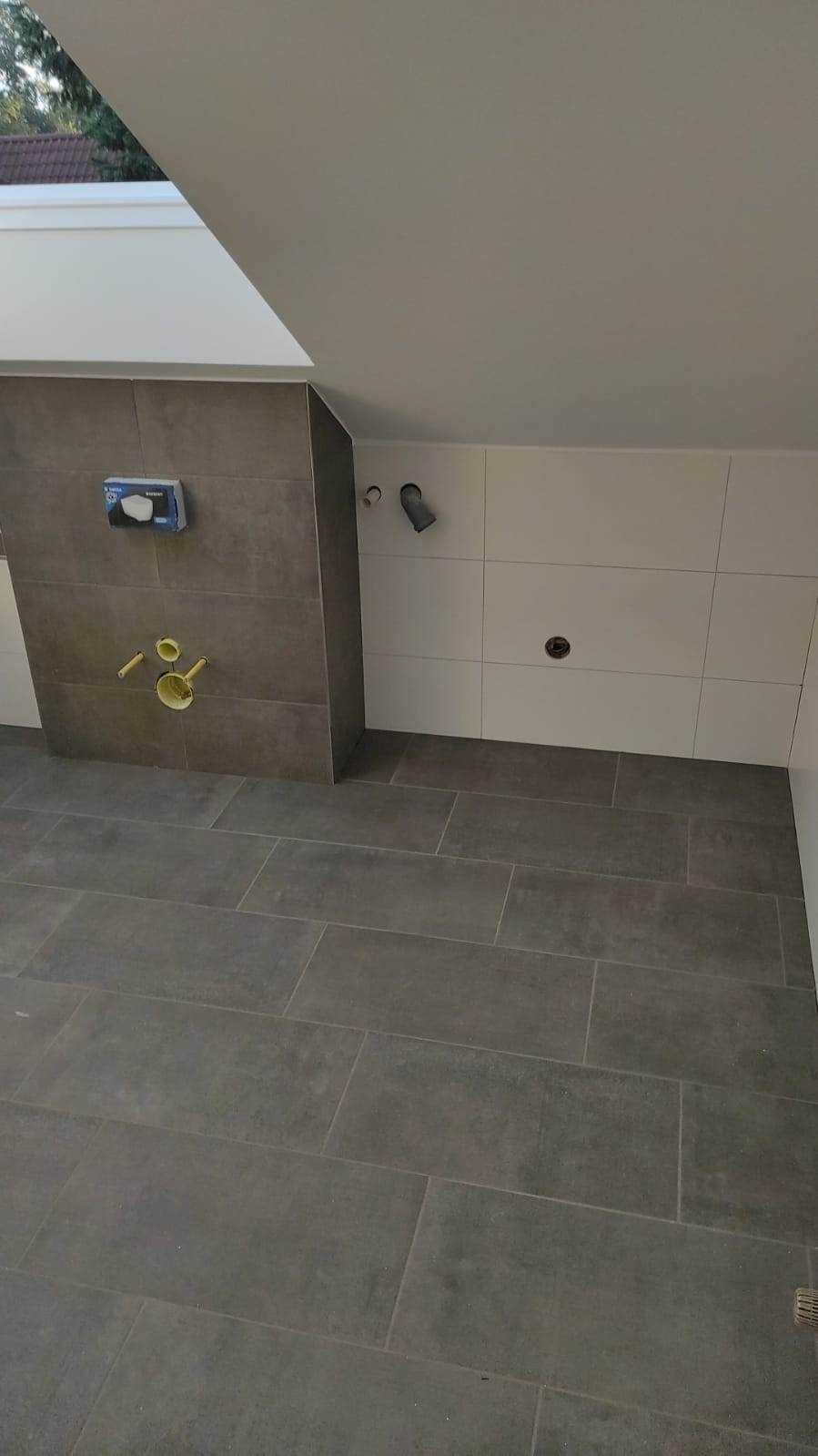 Wnętrze łazienki w trakcie wykańczania, widoczne szare płytki podłogowe, białe płytki ścienne, zabudowa stelaża podtynkowego z szarych płytek oraz wystające rury instalacyjne.