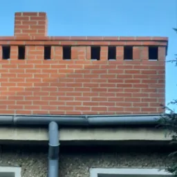 Murowany komin z czerwonej cegły z otworami wentylacyjnymi, zamontowany na dachu z rynną, widoczny fragment elewacji budynku z oknem.