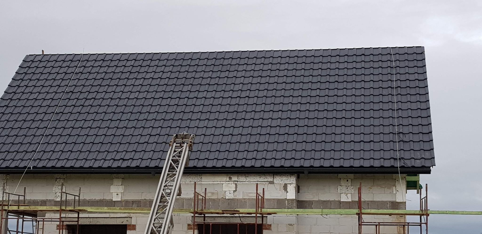 Nowy, grafitowy dach z metalowej dachówki, widoczny fragment rusztowania i drabina przystawiona do dachu, surowy stan elewacji budynku.