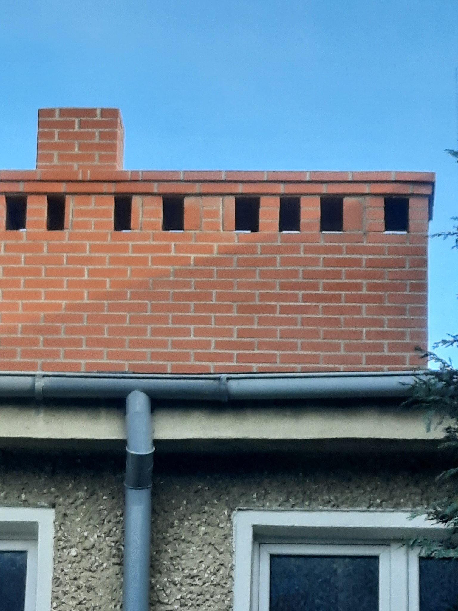 Murowany komin z czerwonej cegły z otworami wentylacyjnymi, zamontowany na dachu z rynną, widoczny fragment elewacji budynku z oknem.