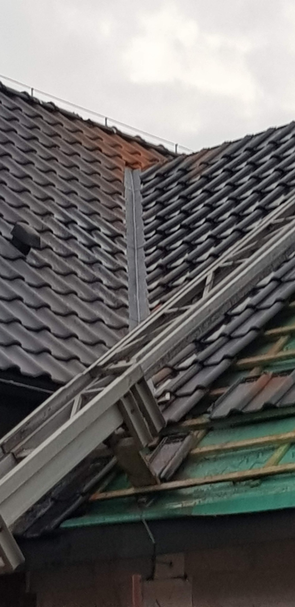 Fragment dachu z szarą blachą okapową i częściowo zdjętymi dachówkami, widoczne elementy konstrukcji drewnianej i drabina dekarska.