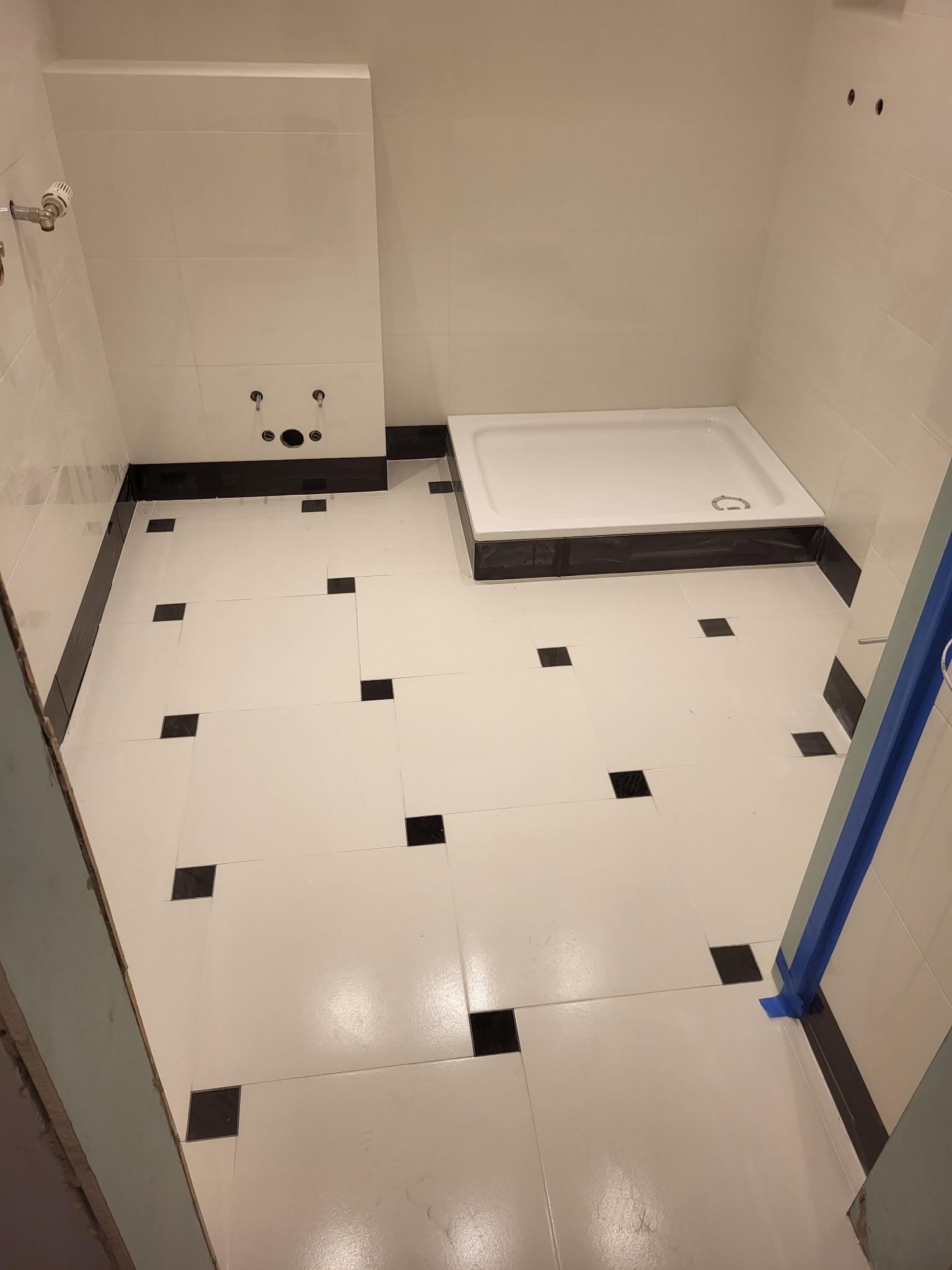 Wnętrze łazienki w trakcie prac wykończeniowych, widoczny brodzik, białe płytki ścienne i podłogowe z czarnymi akcentami, oraz przygotowane miejsce na armaturę.