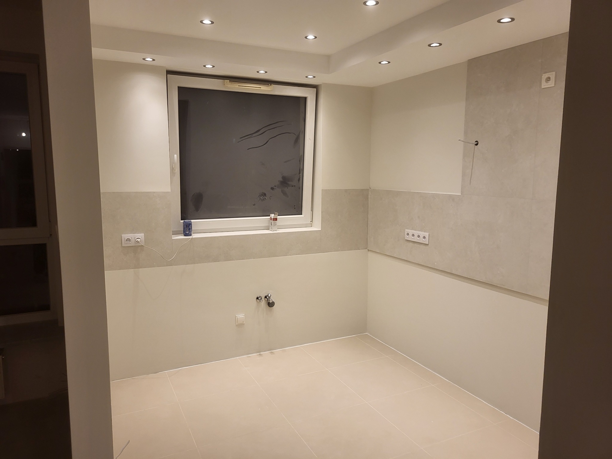 Nowo wykończona, jasna łazienka z beżowymi płytkami podłogowymi i częściowo na ścianach, widoczne punkty oświetleniowe w suficie, instalacja wodna i gniazdka elektryczne, okno zaklejone folią...