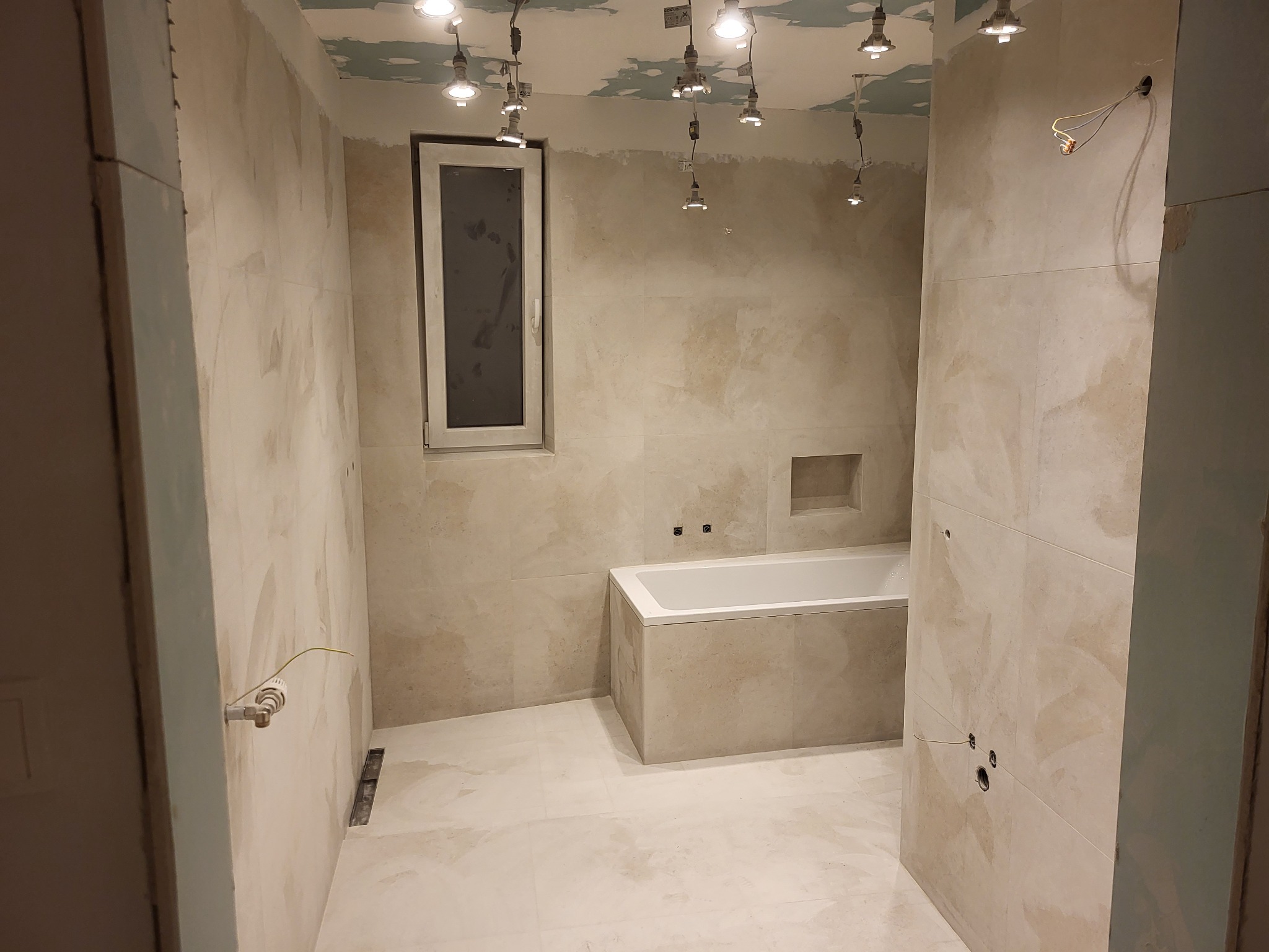 Łazienka w trakcie remontu: ułożone jasne płytki imitujące kamień na ścianach i podłodze, zabudowana wanna, okno, sufit z instalacją oświetleniową przed montażem lamp.