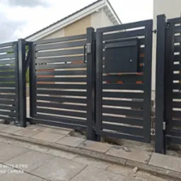 Nowoczesna, pozioma brama wjazdowa z czarnych profili stalowych z drewnianymi akcentami, wyposażona w skrzynkę na listy i panel numeryczny, na tle domu jednorodzinnego.