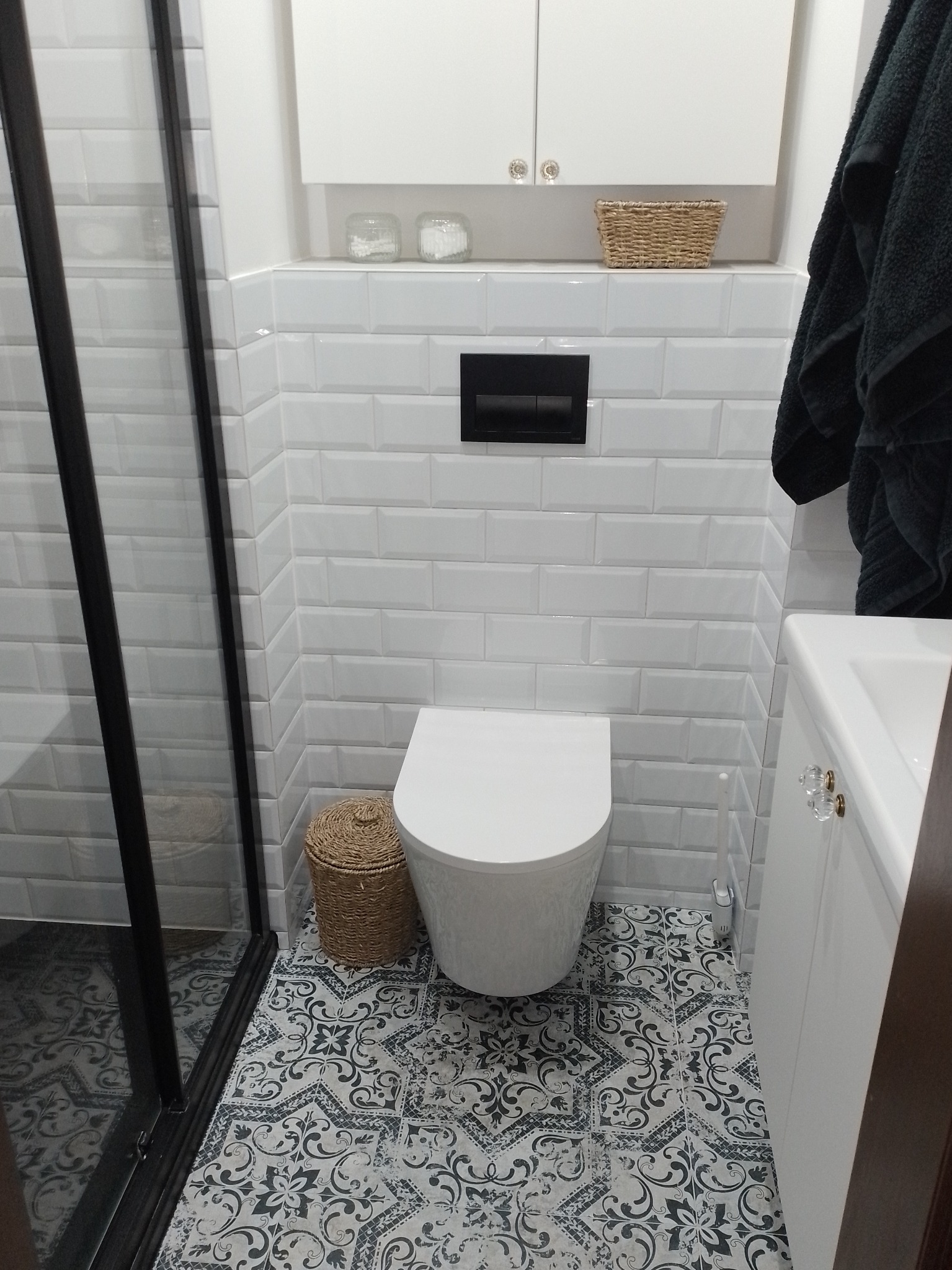 Nowoczesna łazienka z białą armaturą, wzorzystą podłogą, oraz białymi płytkami imitującymi cegłę na ścianach. Widoczna kabina prysznicowa z czarnymi ramami, biała szafka z ozdobnymi uchwytami...