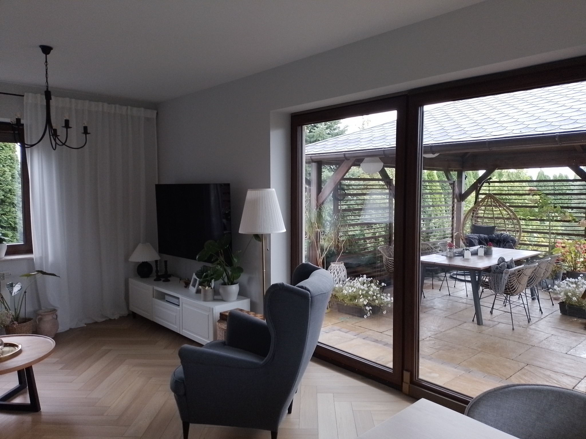 Salon w Jazowsku z parkietem ułożonym w jodełkę, widok na zadaszony taras przez duże okno przesuwne, szary fotel uszak.