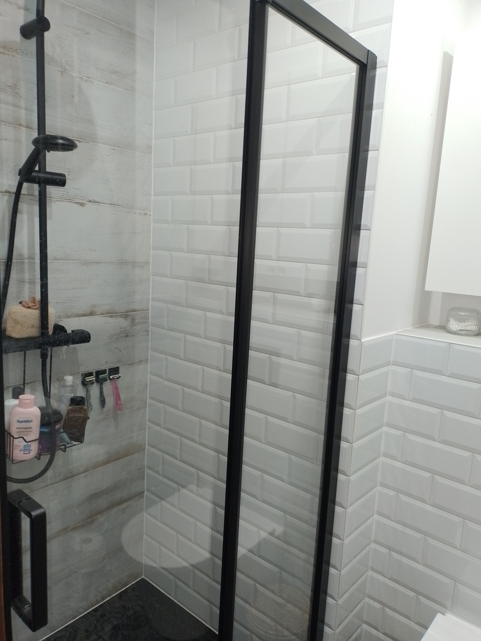 Nowoczesna kabina prysznicowa z czarną ramą i białymi płytkami typu metro, widoczne kosmetyki na półce pod prysznicem.