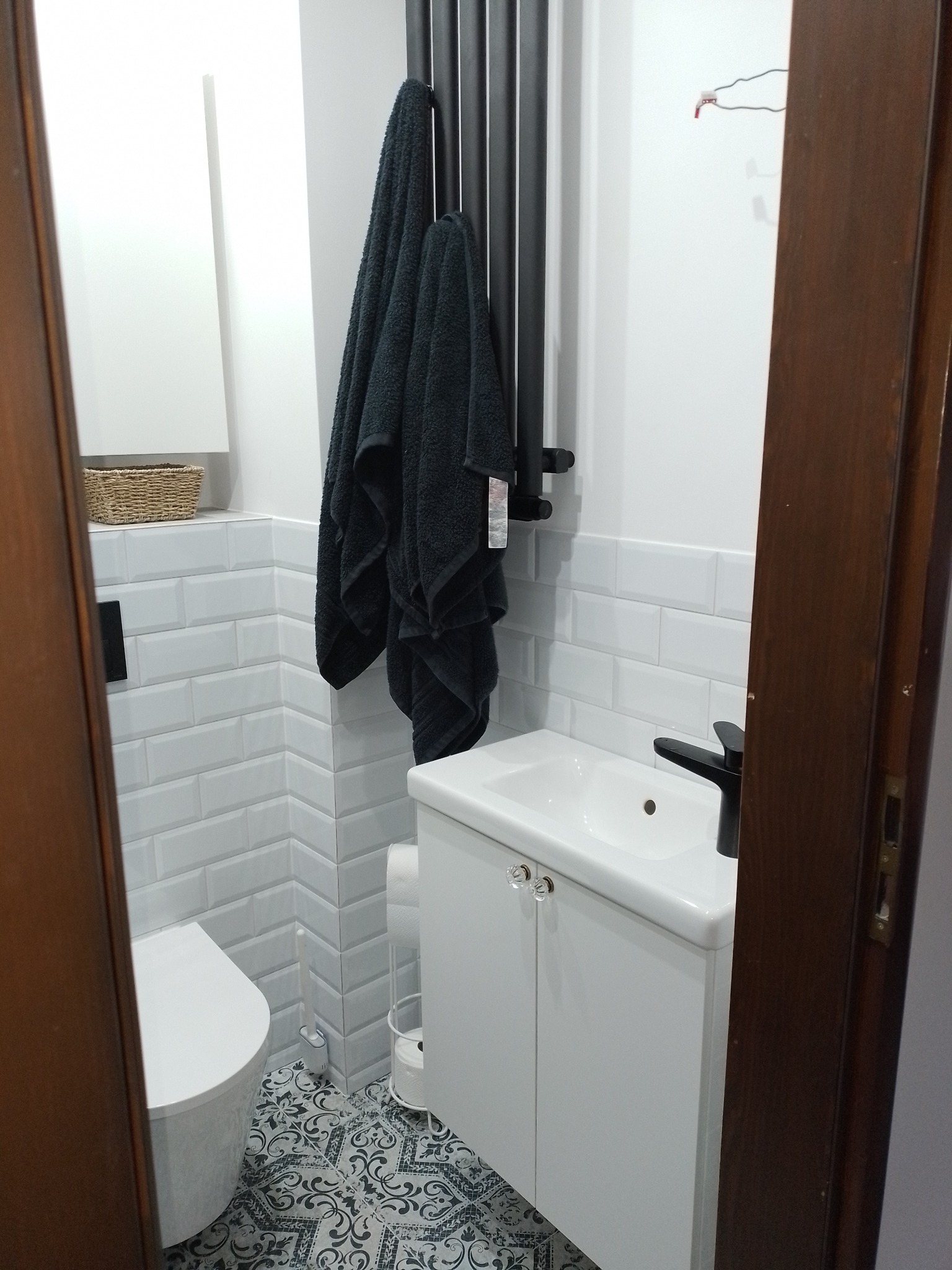 Wykończona łazienka z białą ceramiką, armaturą w kolorze czarnym i wzorzystą podłogą. Na grzejniku wisi czarny ręcznik.