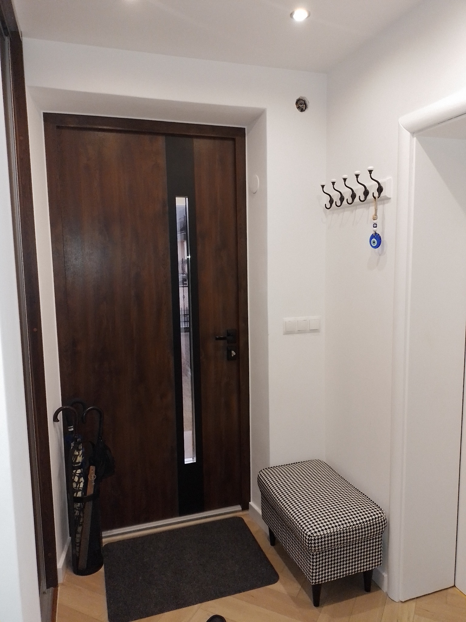 Nowoczesny przedpokój z drewnianymi drzwiami z czarnym paskiem, wieszakiem na ubrania z amuletem Nazar i pufą w pepitkę.