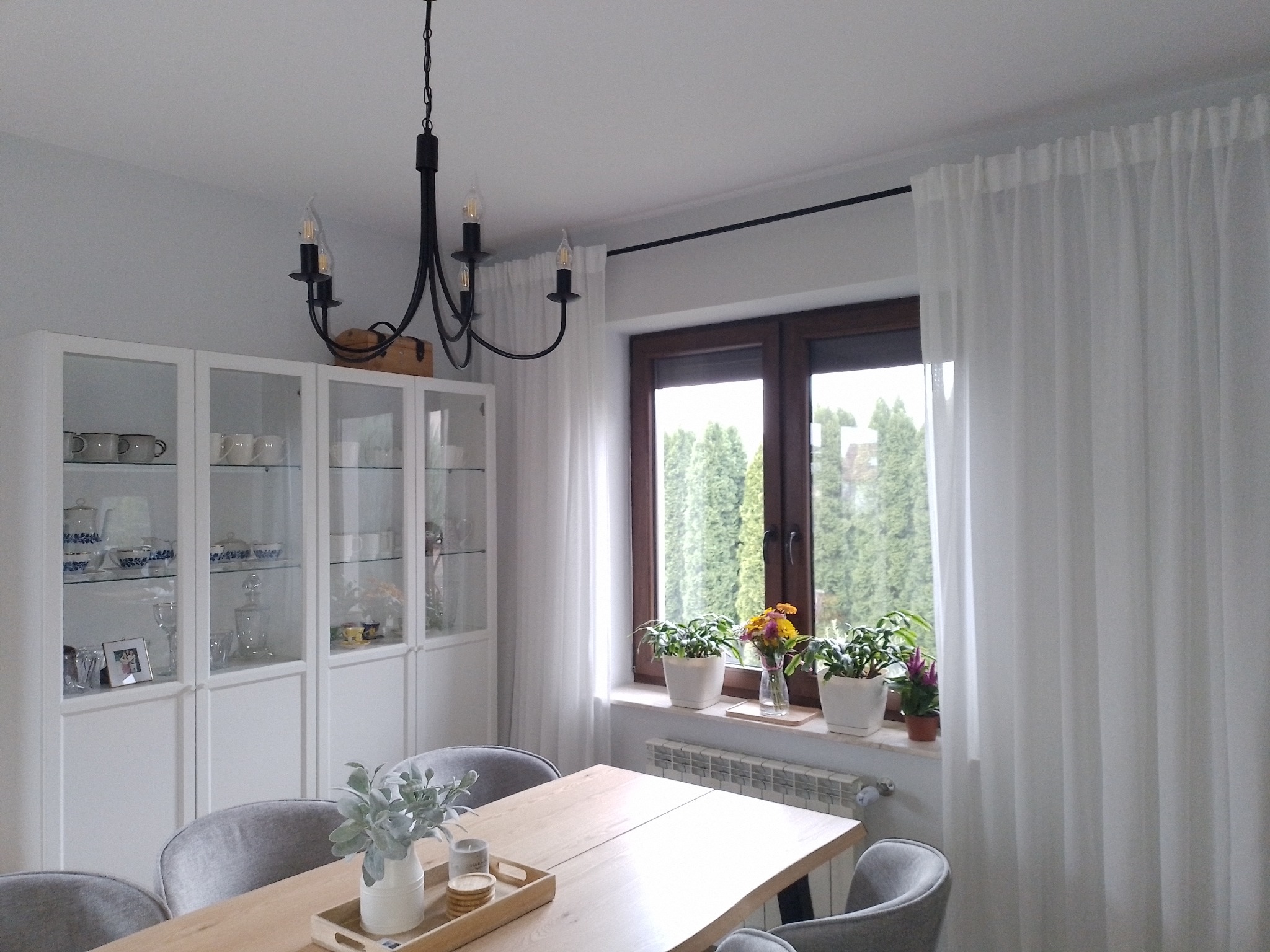 Jasne wnętrze jadalni z drewnianym stołem, szarymi krzesłami, przeszkloną białą witryną, żyrandolem i oknem z białymi firanami oraz roślinami doniczkowymi.