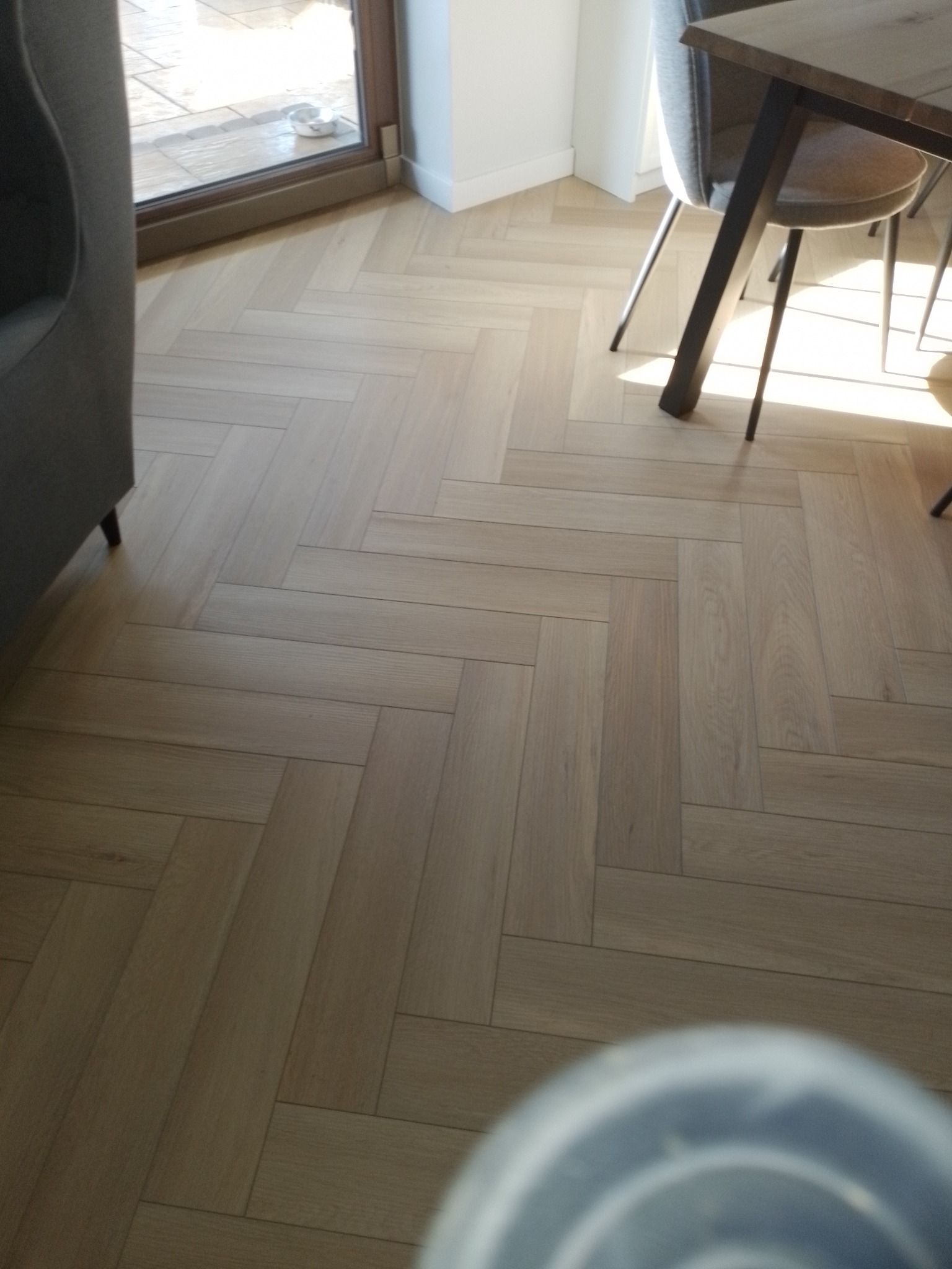 Jasne panele ułożone w jodełkę francuską, widoczne fragmenty mebli: szara sofa i krzesło przy drewnianym stole, naturalne światło z okna