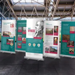 Zestaw roll-upów prezentujących informacje o Józefie Wybickim, w neutralnym wnętrzu z przeszklonymi ścianami i metalową konstrukcją dachu.