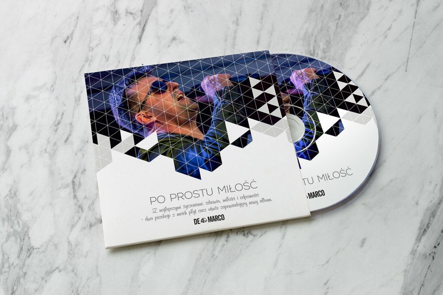 Płyta CD w okładce z geometrycznym wzorem i zdjęciem muzyka śpiewającego do mikrofonu, leżąca na marmurowym tle.