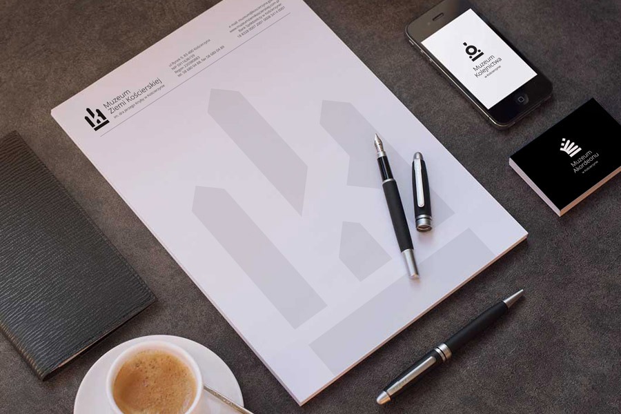 Papier firmowy z logo Muzeum Ziemi Kościerskiej, wizytówka z logo Muzeum Akordeonu, smartfon z logo Muzeum Kolejnictwa, filiżanka kawy i dwa pióra na ciemnym tle.