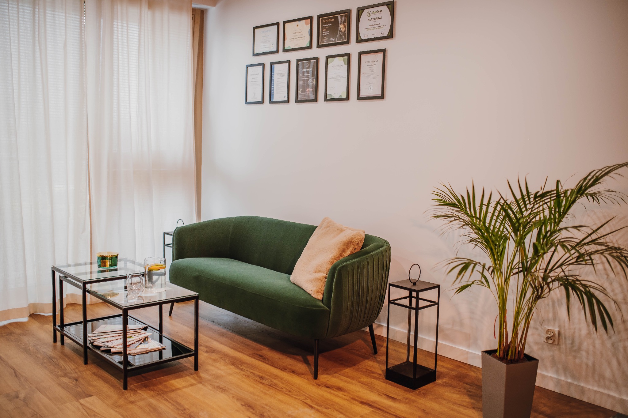 Wnętrze gabinetu z zieloną sofą, stolikiem kawowym z napojami i certyfikatami na ścianie.