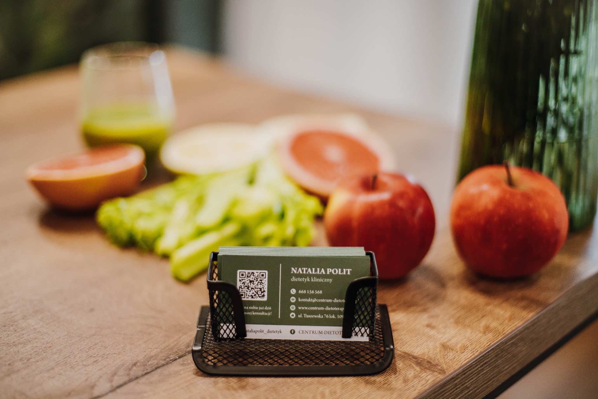 Wizytówka dietetyka klinicznego Natalia Polit w metalowym organizerze na biurku, obok szklanki zielonego soku, grejpfruta, sałaty i jabłek.