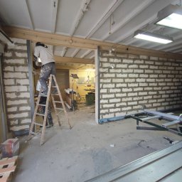 Tomasz Neumann - Pracownik na drabinie maluje ścianę z bloczków betonowych w surowym wnętrzu warsztatu, widoczny fragment piły stołowej i inne narzędzia.