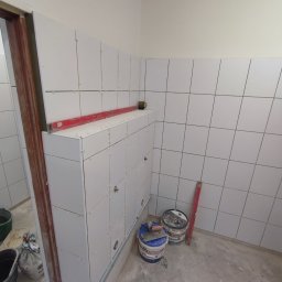 Tomasz Neumann - Białe płytki ceramiczne ułożone na ścianach i zabudowie w łazience, z widocznymi krzyżykami dystansowymi i poziomnicą, obok stoją wiadra z zaprawą i narzędzia do układania płytek.
