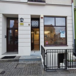 Tomasz Neumann - Odnowiona elewacja budynku z jasną fasadą, ciemnymi ramami okiennymi i drzwiami wejściowymi, widok na wnętrze przez otwarte drzwi, numer budynku 24 nad wejściem.