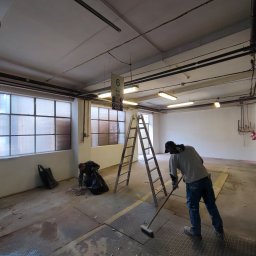 Tomasz Neumann - Dwóch pracowników sprząta podłogę po remoncie w przestronnym pomieszczeniu z dużymi oknami i drabina na środku, jeden zbiera gruz do czarnego worka, drugi zamiata podłogę szczotką.