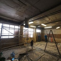 Tomasz Neumann - Wnętrze w trakcie remontu, zabezpieczona folią podłoga, rusztowania przy oknach, widoczni trzej mężczyźni w tle, surowy betonowy sufit z lampami.
