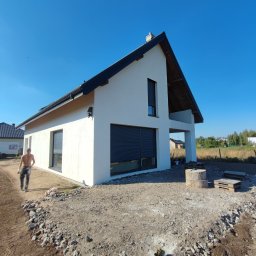 Tomasz Neumann - Nowoczesny dom jednorodzinny z białą elewacją i czarnymi ramami okiennymi, widoczne niedokończone prace ziemne wokół budynku i mężczyzna w oddali.