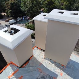 Tomasz Neumann - Trzy pomalowane kominy na dachu, zabezpieczone folią i taśmą malarską, widok z góry na tle drzew i ulicy.