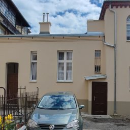 Tomasz Neumann - Odnowiona elewacja budynku z beżową fasadą, widoczne okna z białymi ramami, brązowe drzwi wejściowe, rynna, fragment dachu z kominem oraz zaparkowany samochód osobowy z odbiciem chmur w szybie.