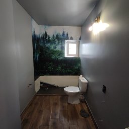 Tomasz Neumann - Wąska łazienka w trakcie remontu: ściana z fototapetą leśną, drewnopodobne panele podłogowe, szara ściana, biała toaleta i okno. Widoczne niedokończone prace przy ścianie.