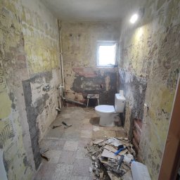 Tomasz Neumann - Łazienka w trakcie demontażu starej glazury i przygotowania do remontu, widoczne odsłonięte cegły, resztki kleju, narzędzia oraz fragmenty płytek na podłodze.