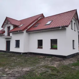 Nowo otynkowany dom jednorodzinny z czerwoną dachówką, w trakcie prac wykończeniowych, z folią ochronną na oknach i widocznymi materiałami budowlanymi na działce.