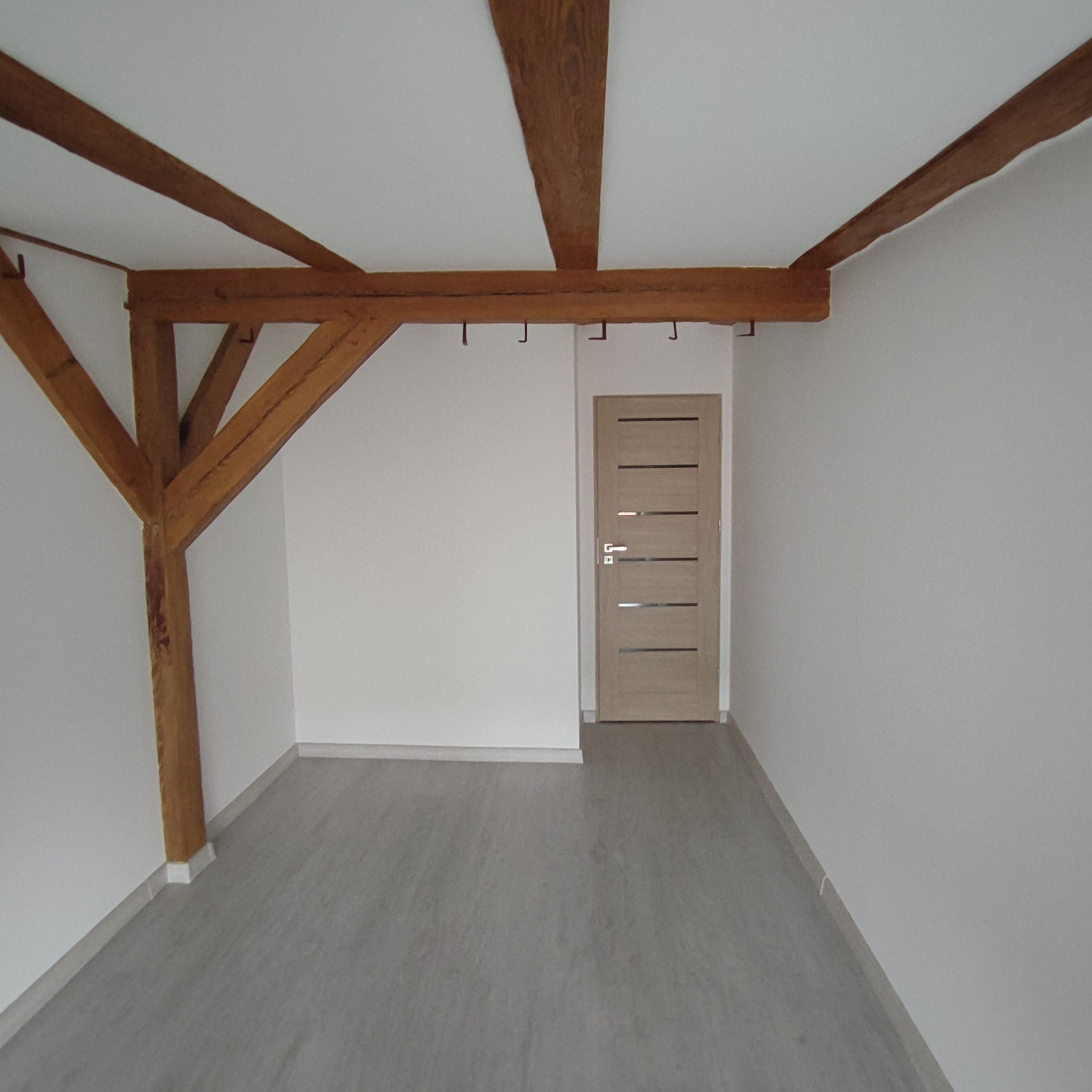 Pusty pokój z drewnianymi belkami stropowymi, szarą podłogą i jasnymi ścianami, widoczna drewniana konstrukcja wspierająca strop, drzwi wewnętrzne z poziomymi przeszkleniami.