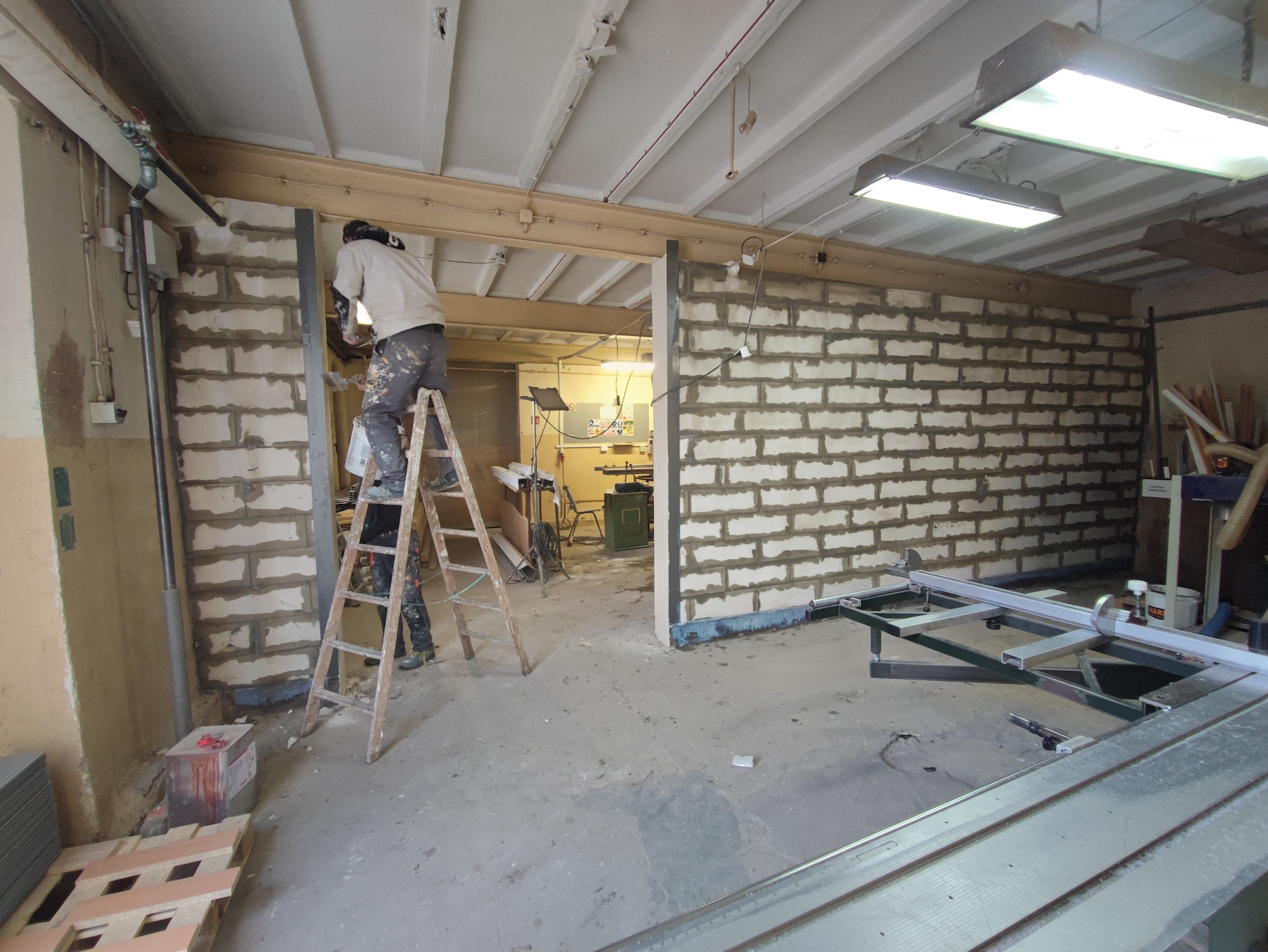 Pracownik na drabinie maluje ścianę z bloczków betonowych w surowym wnętrzu warsztatu, widoczny fragment piły stołowej i inne narzędzia.