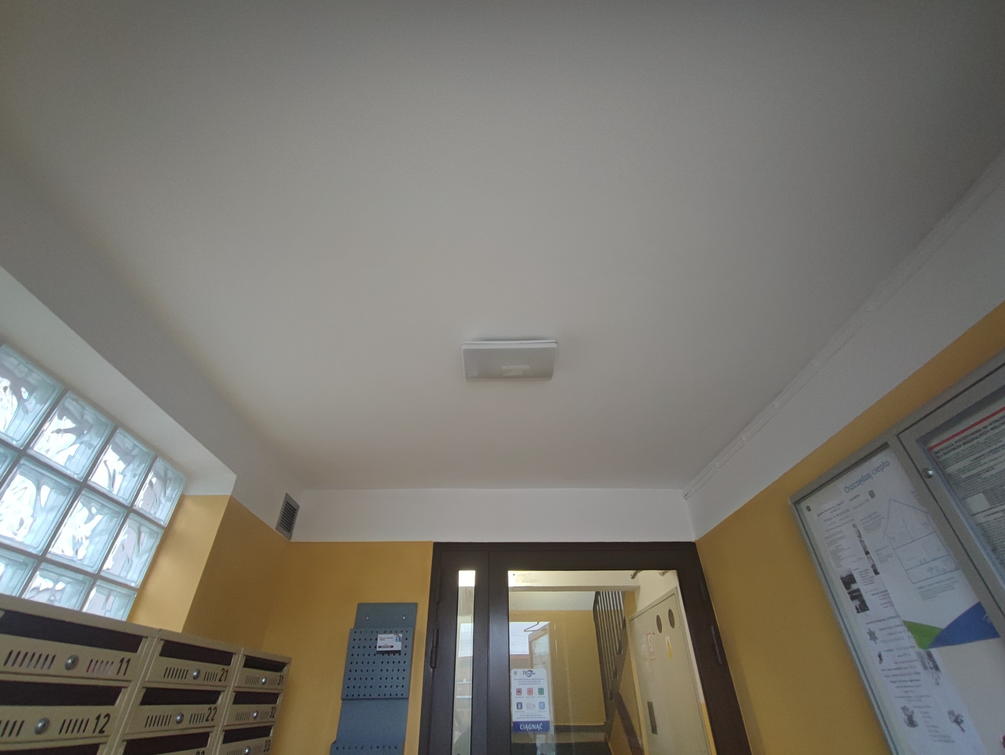 Wnętrze klatki schodowej z nowo pomalowanym sufitem na biało, widoczne skrzynki pocztowe, okno z luksferami i tablica informacyjna na ścianie.