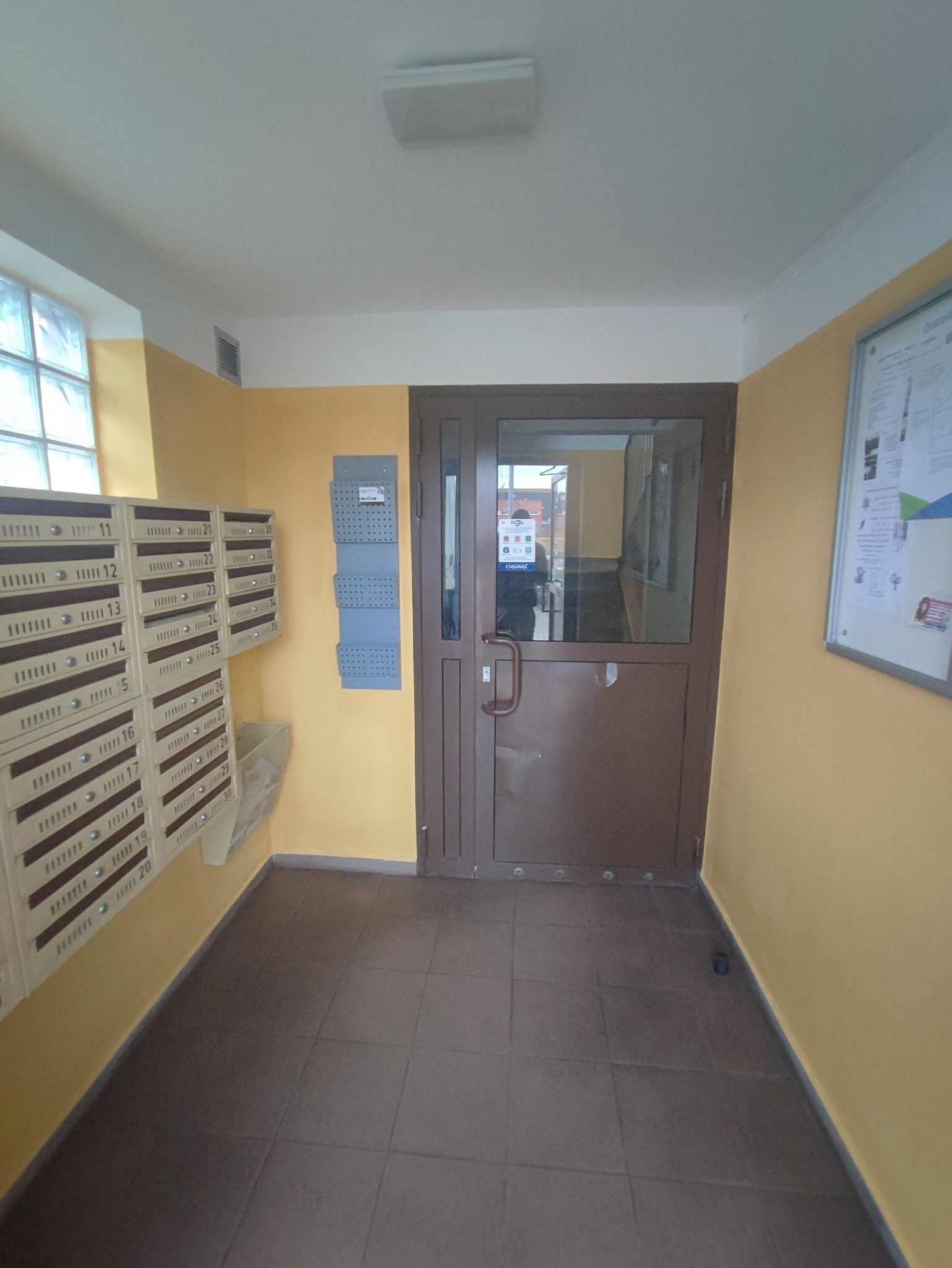 Korytarz wejściowy do budynku wielorodzinnego z rzędami skrzynek pocztowych po lewej stronie i brązowymi drzwiami wejściowymi na wprost, ściany pomalowane na żółto.