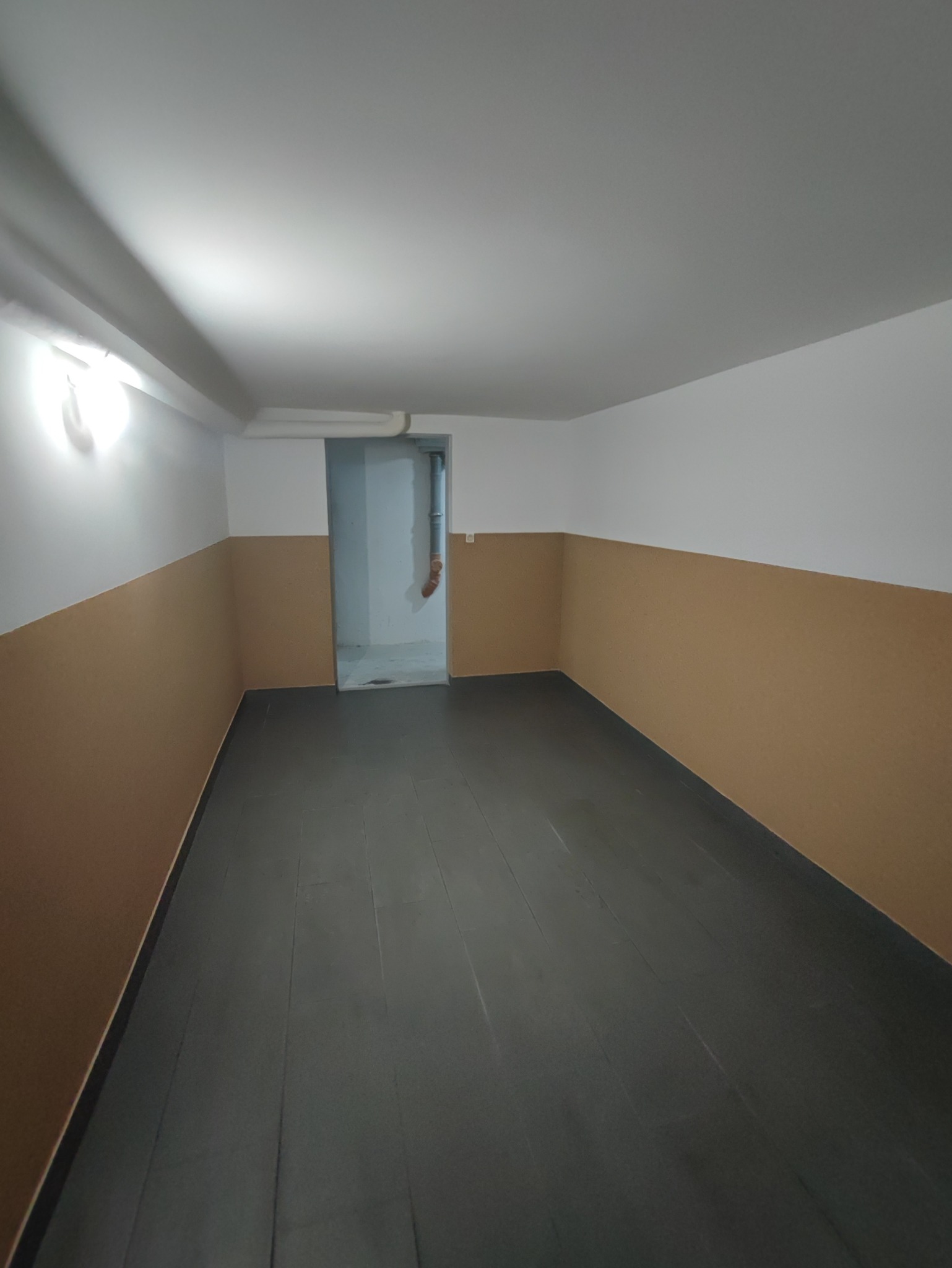 Pusta piwnica w Toruniu po remoncie, z pomalowanymi ścianami do połowy na brązowo i szarą podłogą, widoczny otwór drzwiowy do innego pomieszczenia.