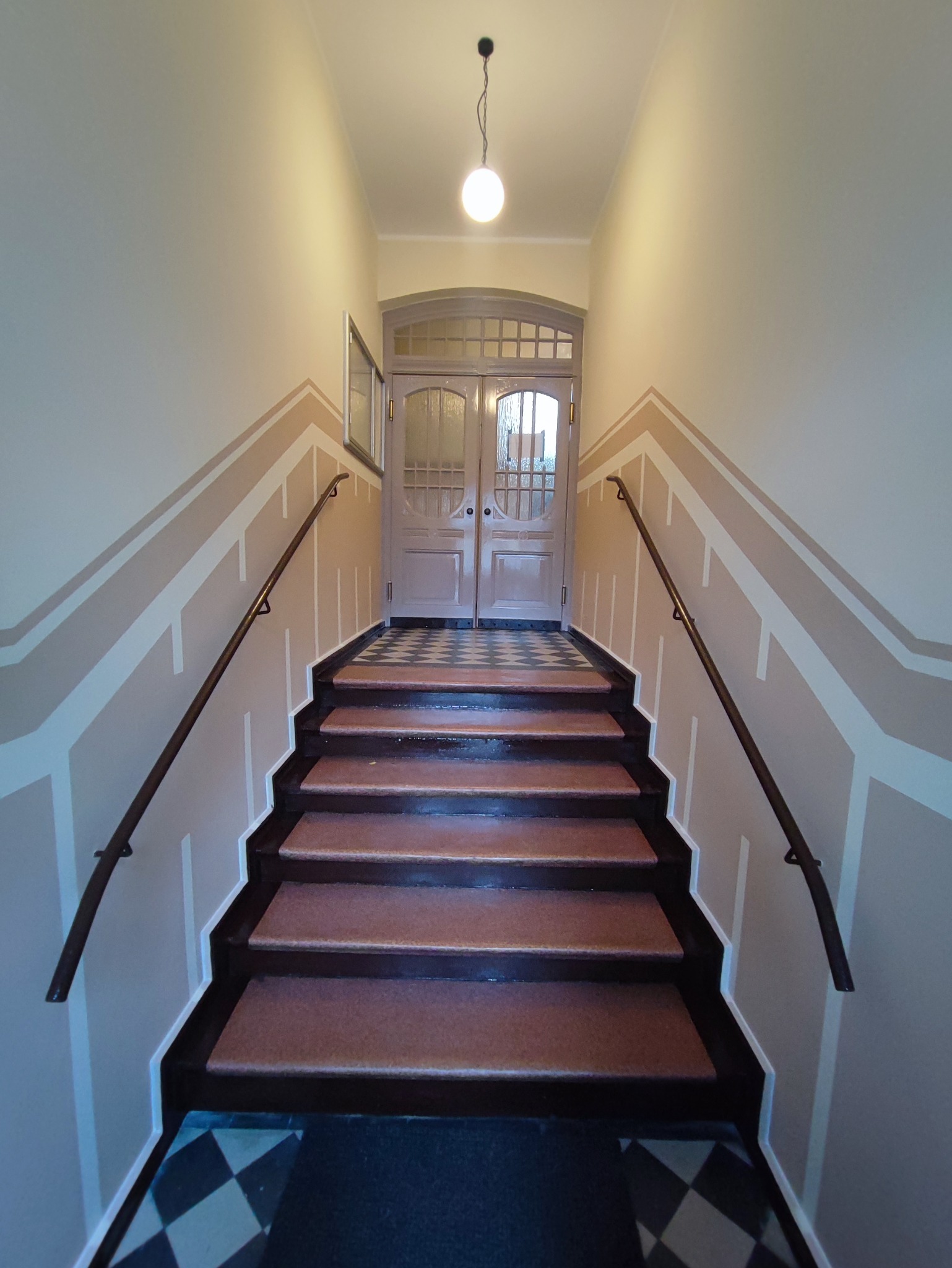 Klatka schodowa z odnowionymi ścianami w geometryczne wzory, prowadząca do dwuskrzydłowych drzwi z przeszklonymi panelami, oświetlona wiszącą lampą.