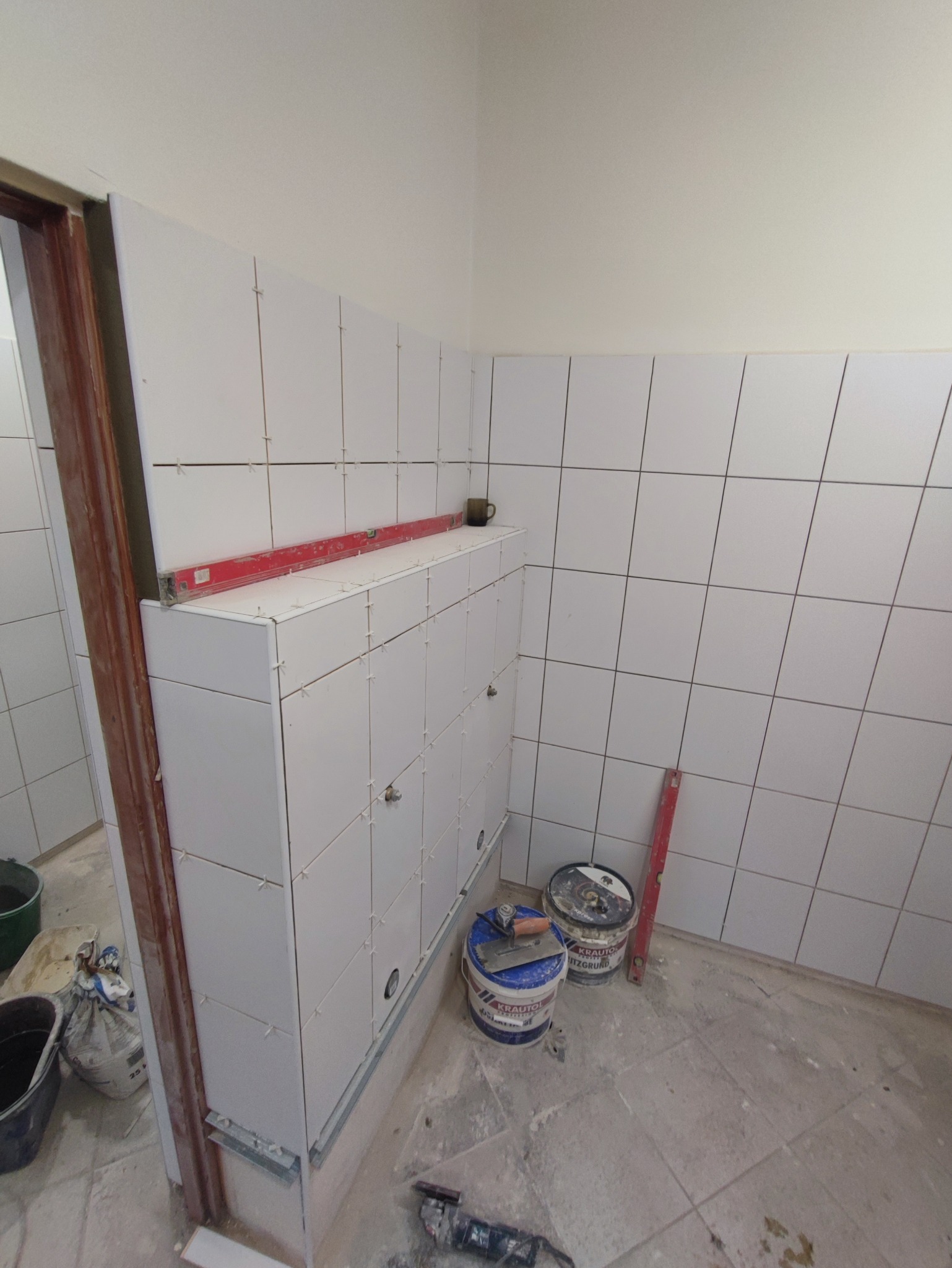 Białe płytki ceramiczne ułożone na ścianach i zabudowie w łazience, z widocznymi krzyżykami dystansowymi i poziomnicą, obok stoją wiadra z zaprawą i narzędzia do układania płytek.